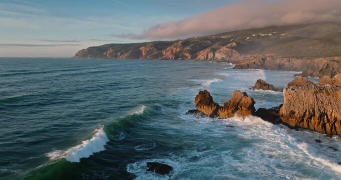 Portuguese coastline revealing the rugged cliffs and dramatic rock formations of Sintra Cascais Natural Park, powerful ocean waves breaking against the shore during a warm sunset. Dramatic landscape