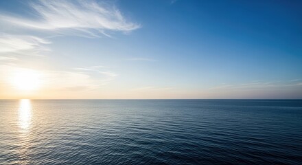 Fototapeta premium The sun rises over a calm ocean, creating a shimmering reflection on the water. Wispy clouds drift across a clear blue sky.