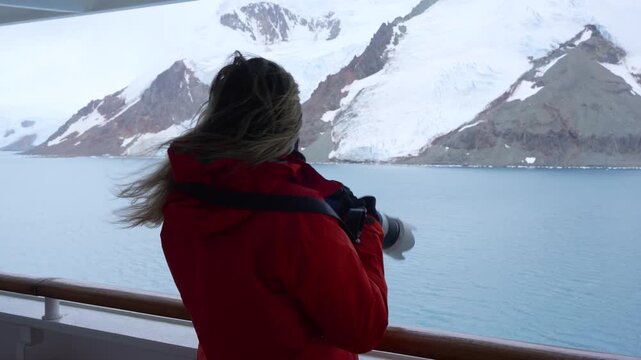 Woman Photographer on Antarctica Cruise Ship Takes Photos with Professional Camera in Orange Red Coat. Wind Blows Hair. Snow Solo Female Travel