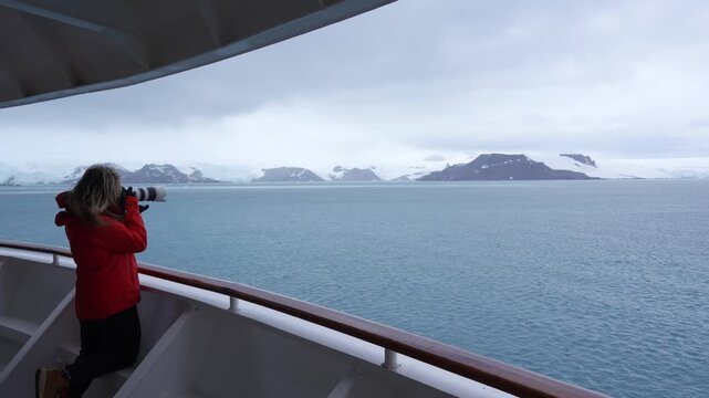 Woman Photographer on Antarctica Cruise Ship Takes Photos Wide Shot Professional Camera in Orange Red Coat. Wind Blows Hair. Snow Solo Female Travel