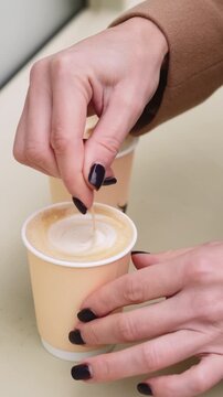 Vertical video. Stirring coffee. Takeaway drink. Female hand in coat with nail polish mixing latte beverage in paper cup with wooden stick.