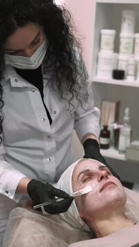 Vertical video. Cream mask. Facial beauty. Esthetician performing professional care applying white product on female skin using brush in salon.