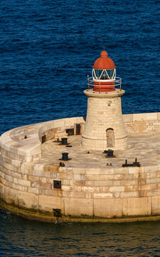 &Aacute;ngulo vertical de un cl&aacute;sico faro mar&iacute;timo con c&uacute;pula roja situado en un muelle de piedra frente a las olas del mar azul oscuro.