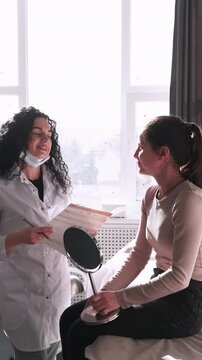 Vertical video. Beauty consultation. Aesthetic clinic. Professional cosmetologist discussing treatment with female patient using mirror at modern workplace.