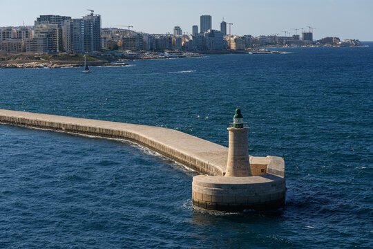 Vista panor&aacute;mica de un faro verde al final de un largo rompeolas de piedra, con el skyline de una moderna ciudad costera al fondo.