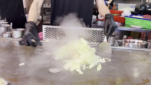 Chef Stir Frying Cabbage On Steaming Teppanyaki Griddle Chef rapidly stir-fries chopped cabbage with two metal spatulas on a steaming teppanyaki griddle in an open restaurant kitchen.