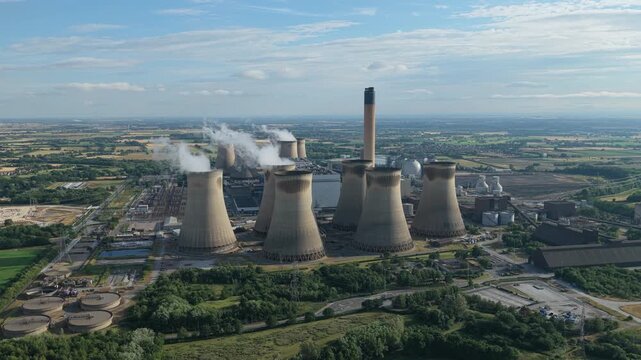Aerial view of a large thermal power station with many cooling towers and a tall smoke stack releases white steam on a sunny day in the countryside.