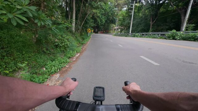 First-person POV of a road cyclist riding along a winding mountain road surrounded by lush tropical forest. The footage captures a smooth cycling experience through scenic curves with chevron warning 