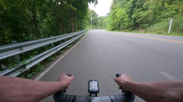 First-person POV footage of a road cyclist riding through a winding mountain road surrounded by lush tropical forest. The clip features oncoming vehicles, roadside guardrails, and curve warning signs,