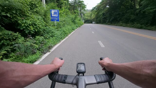 First-person POV footage of a road cyclist riding along a quiet mountain road surrounded by lush tropical forest. The smooth asphalt road, peaceful natural scenery, and distant oncoming vehicle create