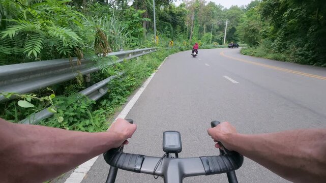 First-person POV footage of a road cyclist riding through a smooth mountain curve surrounded by lush tropical forest. The quiet winding road, roadside guardrails, and chevron warning signs create an i
