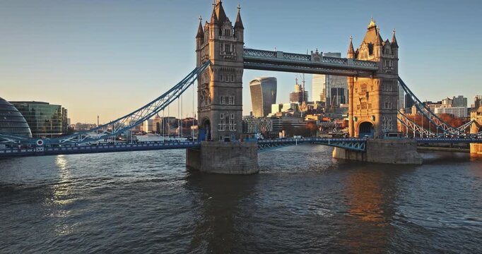 Tower Bridge, an iconic London landmark, spanning the River Thames with the modern city skyline and Walkie Talkie building in the background, illuminated by warm sunset light. Aerial drone flight