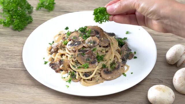 Close-up of a hand garnishing creamy mushroom spaghetti with fresh parsley on a white plate. Homemade Italian pasta with saut&eacute;ed mushrooms in a rich cream sauce, captured in a bright kitchen setting. 