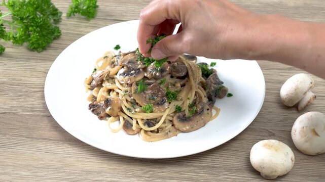 Close-up of a hand garnishing creamy mushroom spaghetti with fresh parsley on a white plate. Homemade Italian pasta with saut&eacute;ed mushrooms in a rich cream sauce, captured in a bright kitchen setting. 