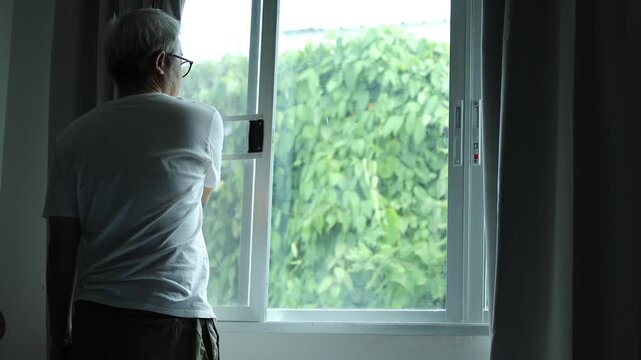 Horizontal 4K back view video of a mature East Asian man in his 50s opening bedroom curtains to look out at green trees and let natural light in