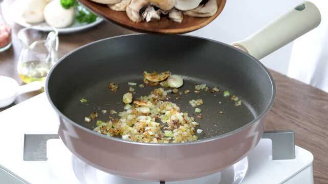 Close-up footage of fresh sliced white button mushrooms being added to a frying pan with saut&eacute;ed onions during home cooking. Healthy vegetarian meal preparation in a modern kitchen with natural light.
