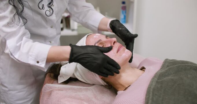 Facial service. Cosmetologist working. Woman professional in black gloves applying white cream moisturizing product in beauty salon.