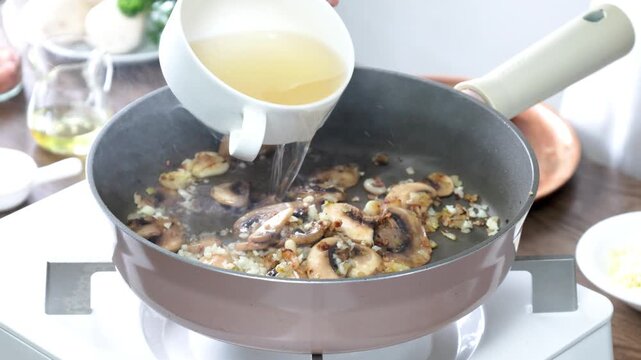 A chef pours chicken broth from a white cup into a non-stick frying pan containing saut&eacute;ed sliced champignon mushrooms, minced garlic, and shallots. with steam clearly rising. In the background, other