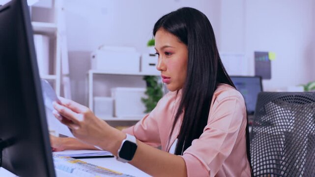 Asian woman office employee reviewing document paperwork at computer desk with concentration, focused business worker in modern workplace with monitor, chair, smartwatch, pink shirt