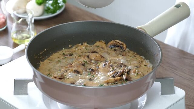 Hand sprinkling grated Parmesan cheese over a creamy mushroom sauce simmering in a frying pan. Homemade Italian-style cream sauce with saut&eacute;ed mushrooms, prepared fresh in a modern kitchen. 