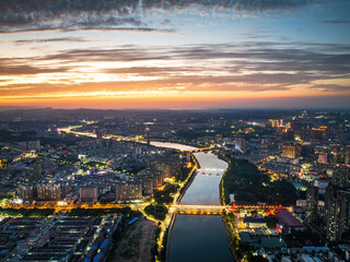 Fototapeta premium Aerial shot of a river running through a lit urban city at dusk, with bridges, buildings and colorful twilight sky.