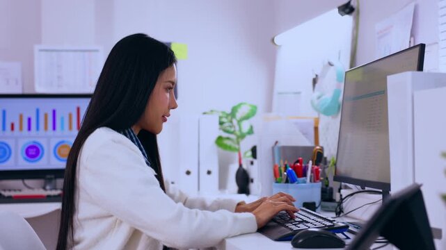 Asian woman typing at computer desk in office workspace, professional business data chart analysis with monitor, keyboard, plant, stationery, modern digital workplace