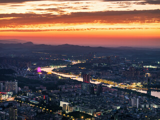 Fototapeta premium A panoramic urban view at dusk, featuring a winding river, illuminated buildings, and a colorful sky with distant mountains.