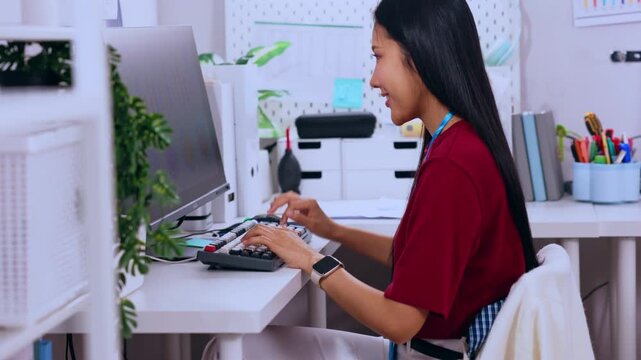 Asian woman using office computer and keyboard at desk with calculator in professional workplace business scene, smiling while typing in bright modern workspace with plant stationery