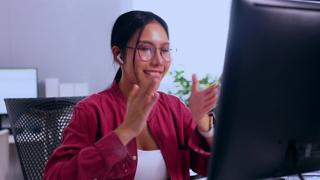 Asian woman in office wearing eyeglass and headset smiling at desk using computer for remote work video call, happy employee in modern workplace communication setup