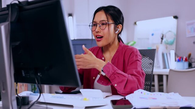 Asian woman office computer eyeglass headset desk document professional business smiling worker using monitor with paper notebook keyboard modern workspace video call remote job