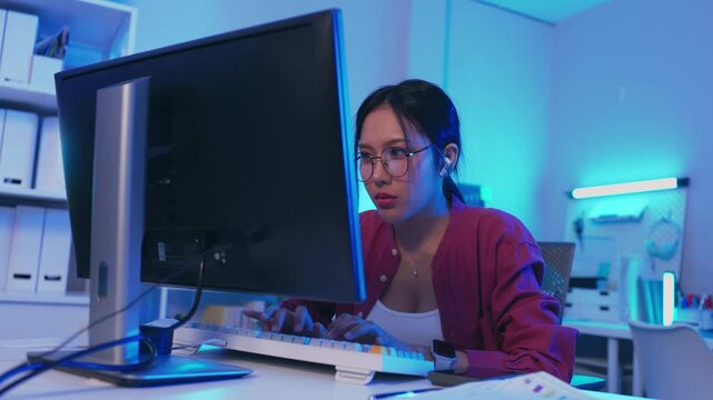 Asian woman typing on keyboard at computer monitor in modern office workspace at night with blue light focused desk documents technology eyeglasses young professional