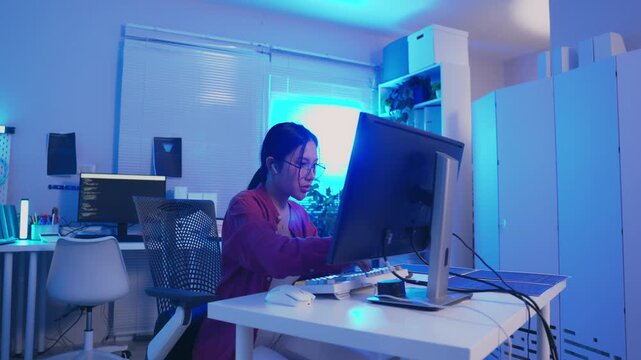Woman computer office desk monitor night blue workspace technology coding focused professional working at home with keyboard mouse cable chair window blind modern room setup