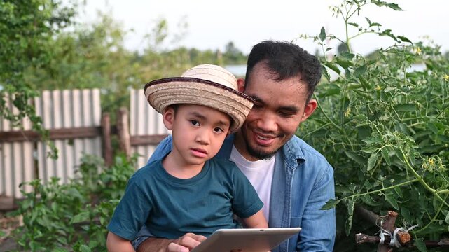 Modern Asian farmer teaching young son agricultural technology using digital tablet in organic garden