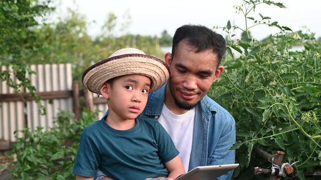 Modern Asian farmer teaching young son agricultural technology using digital tablet in organic garden
