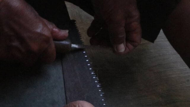Close-up footage of a carpenter's hands carefully sharpening the teeth of a traditional hand saw using a metal file in a rural village in Java, Indonesia. 