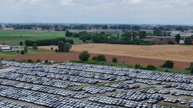 Thousands of passenger vehicles occupy organized inventory rows bordering cultivated farmland, rural shelterbelts and agricultural fields, illustrating large-scale automotive logistics and vehicle dis