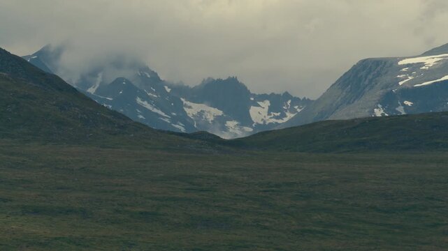 Jagged ridgeline with patchy summer snow looms behind a broad grassy highland under a heavy sky.