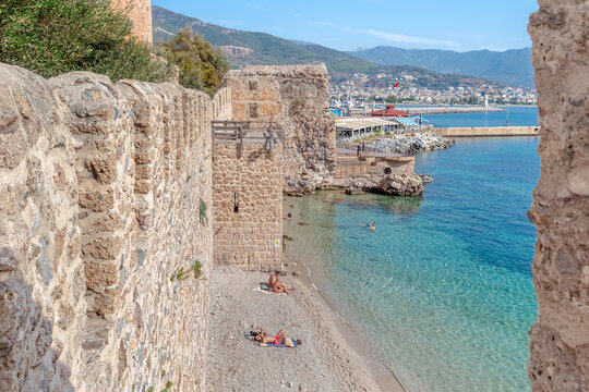 Scenic view of Tersane Beach and Alanya harbor framed by ancient fortress walls. Tourists sunbathing on a pebbly shore near the Red Tower. Alanya, Turkey - October 25, 2020