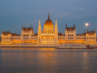 Obraz premium Illuminated Hungarian Parliament Building along the Danube River at blue hour, with warm golden lights reflecting on the water and the moon rising over Budapest, Hungary