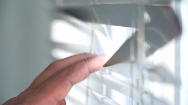 A hand gently opens window blinds, allowing natural sunlight into a modern home interior. Close-up lifestyle footage highlighting daylight, comfort, privacy control, and home living.