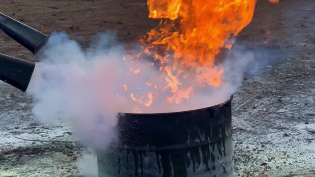 Fire extinguisher spraying white smoke to put out flames in metal bin. Fire safety, emergency response, and firefighting concept footage.
