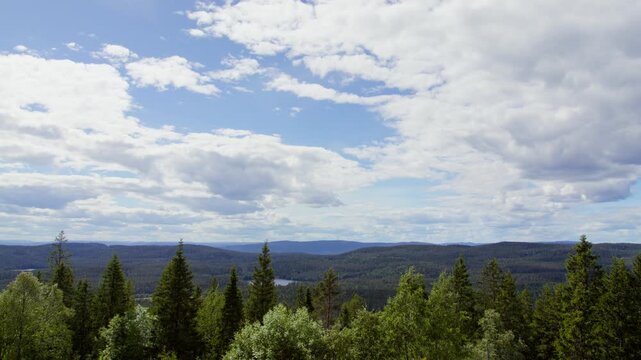 Evergreen and broadleaf trees stretch across a wide valley with layered ridges and a small lake beneath a bright, cloud-filled sky.