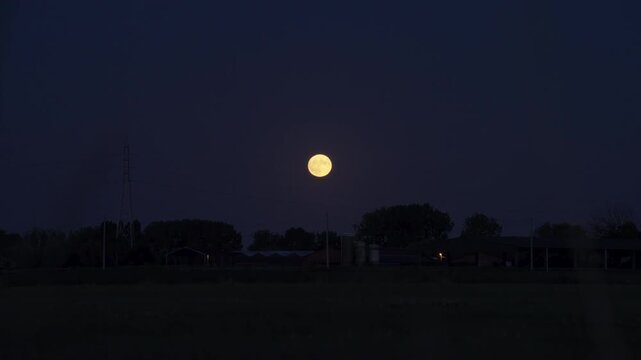Strawberry Moon on a serene night scene featuring a full moon rising over a rural landscape with silhouetted trees and buildings visible dynamic movement atmospheric lighting