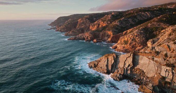 Sintra Cascais Natural Park dramatic ocean waves crashing against rugged cliffs and rock formations along the wild coastline of Portugal, glowing in the warm light of a sunset. Aerial drone flight