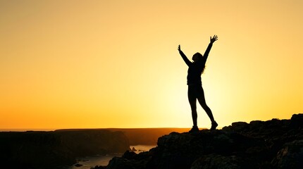 Fototapeta premium Full body silhouette of a woman hiker standing on a rocky cliff edge with arms raised toward a bright yellow sunset sky over a coastal landscape and ocean horizon.