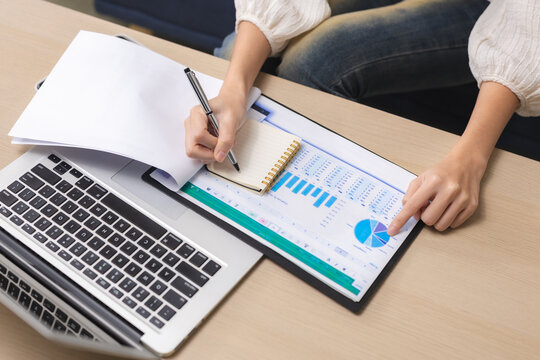 Business person writing in notebook while reviewing chart and graph on desk next to laptop performing careful data analysis for future corporate strategy and financial planning
