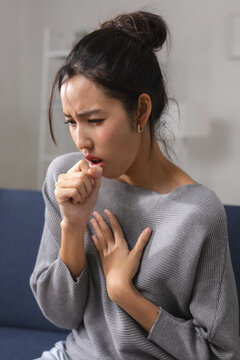 Young asian woman suffering from severe sick cough feeling highly distressed with intense chest pain sitting on sofa looking thoroughly unwell and ill from serious respiratory disease