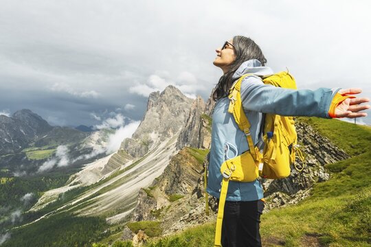 Happy female traveler yellow backpack standing outstretched arms mountain ridge line Seceda South Tyrol Dolomites Italy looking up feeling free peaceful active lifestyle outdoor vacation