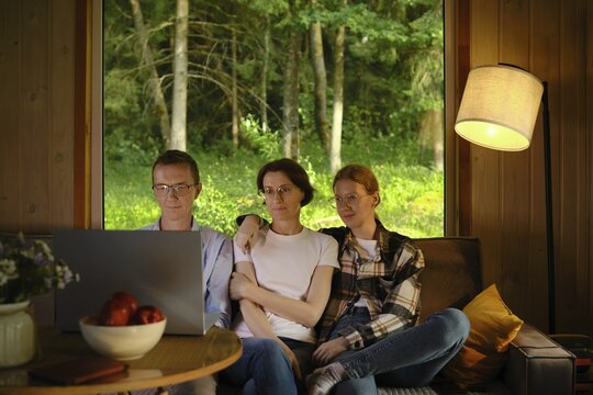 Family sit closely on couch in wooden cabin and watching series on laptop enjoying time together. Concept of advantage of using online streaming platform subscription