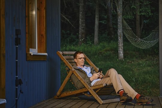 Middle-aged man sits on wooden chair outside wooden cabin in forest. He uses smartphone and enjoys quiet time. Trees surround space and light is soft. A hammock hangs nearby for resting